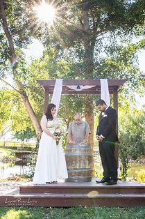 Bride and groom at wedding ceremony under a wooden arbor, sunlight beaming through the trees.