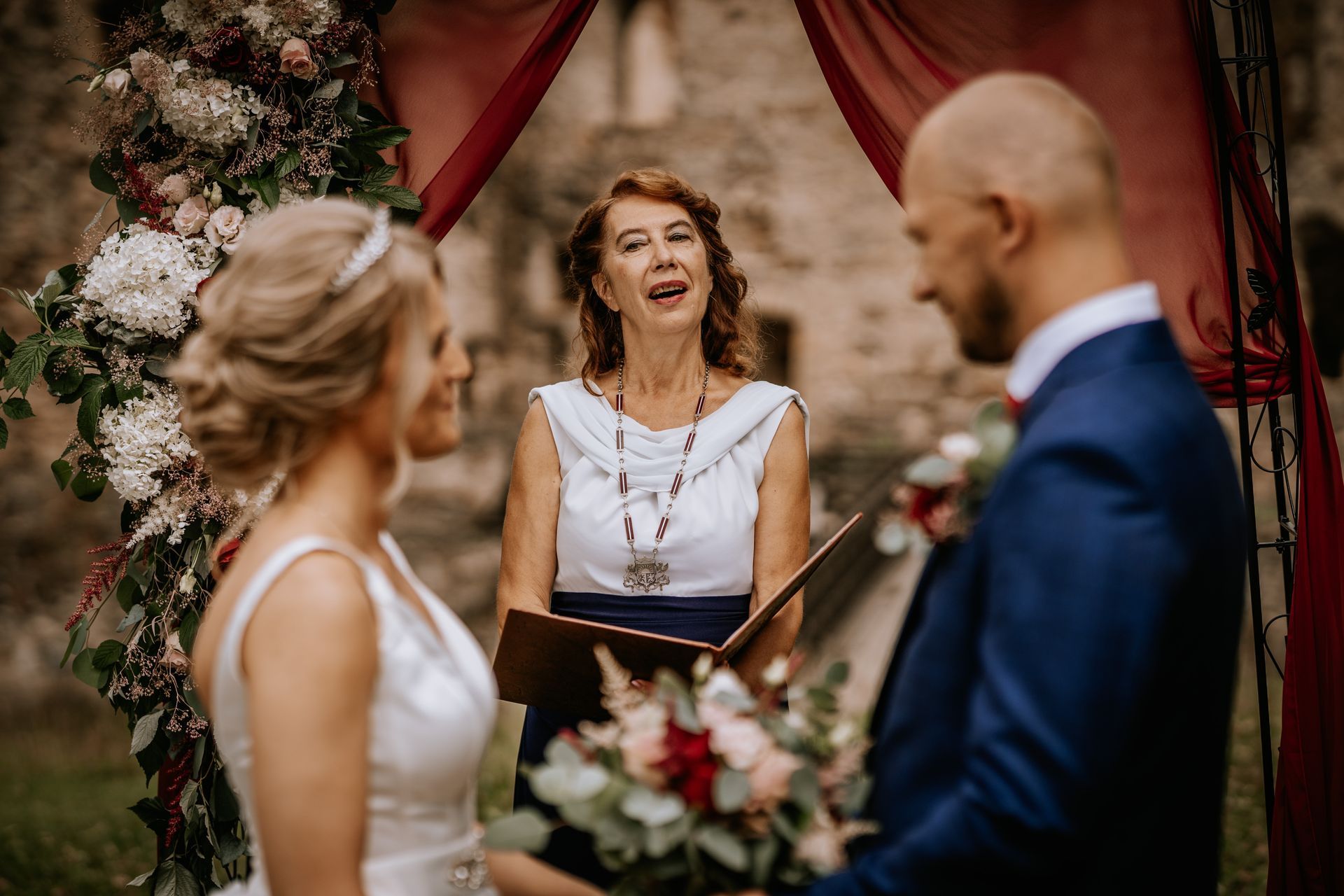 Wedding officiant speaking at outdoor ceremony with couple in foreground and floral arch backdrop