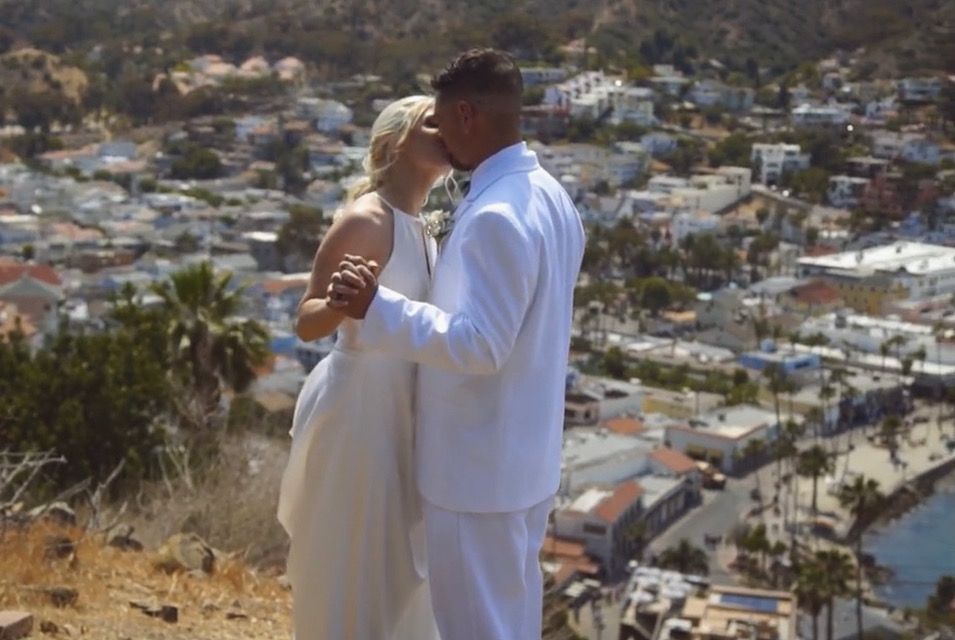 Wedding reception with round tables set with burgundy linens and white chairs, illuminated by purple and gold lighting.