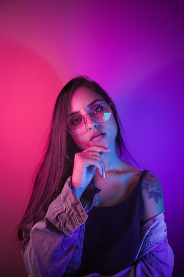 Woman with sunglasses, hand on chin, under pink and purple neon lights.