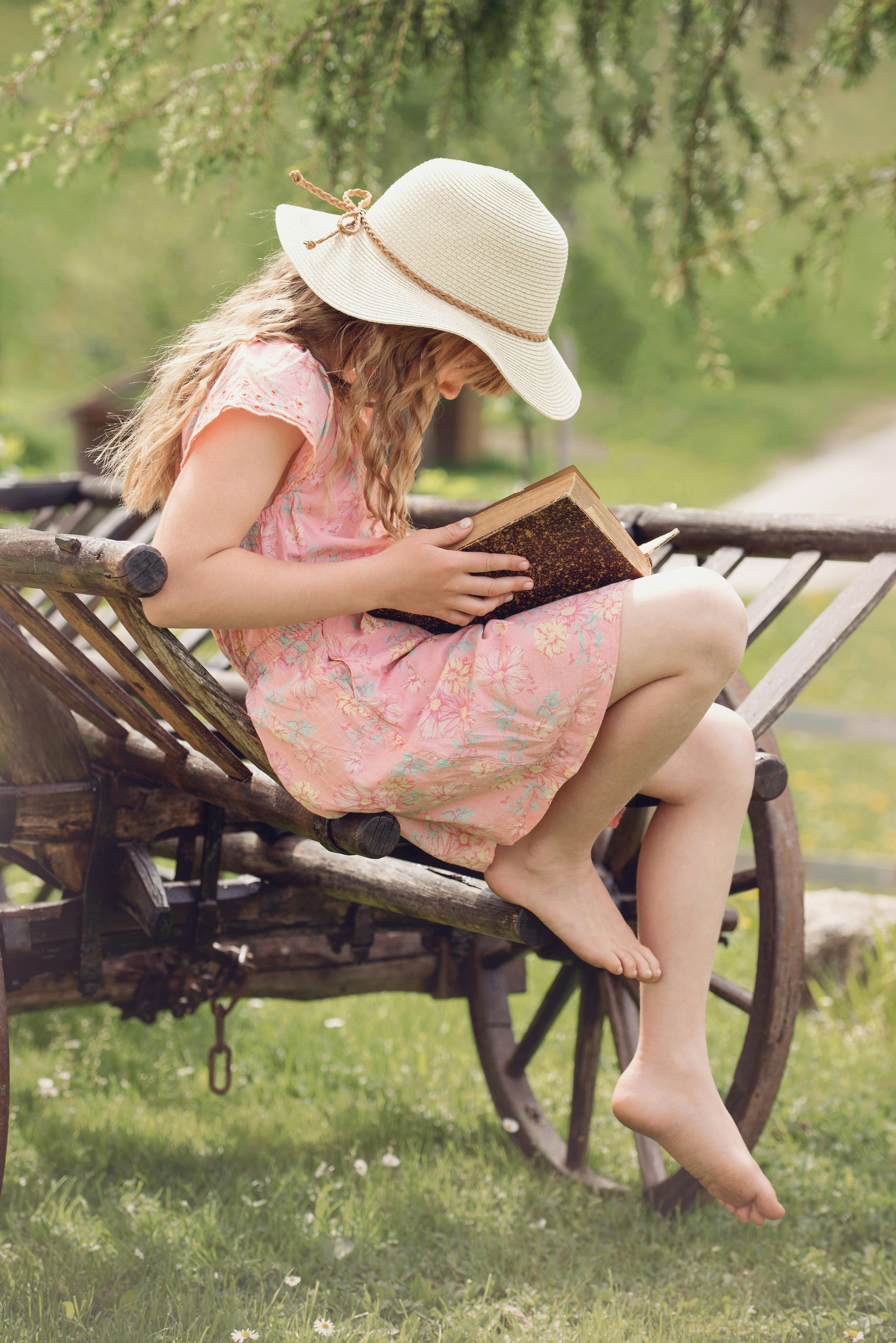 Girl in straw hat reads a book while sitting on a wooden cart in a grassy outdoor setting.
