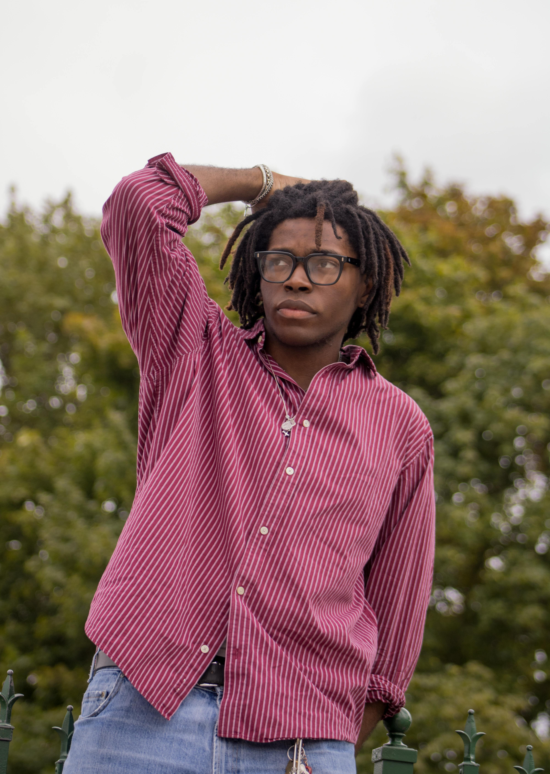 A young person with dreadlocks and glasses in a maroon patterned shirt poses outdoors.