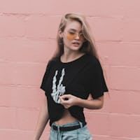 Woman with blonde hair wearing a black tee with a skeleton hand print, sunglasses, against a pink brick wall.