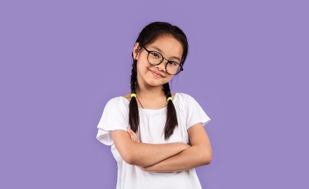 Girl with pigtails, glasses, and crossed arms smiles against a purple background.