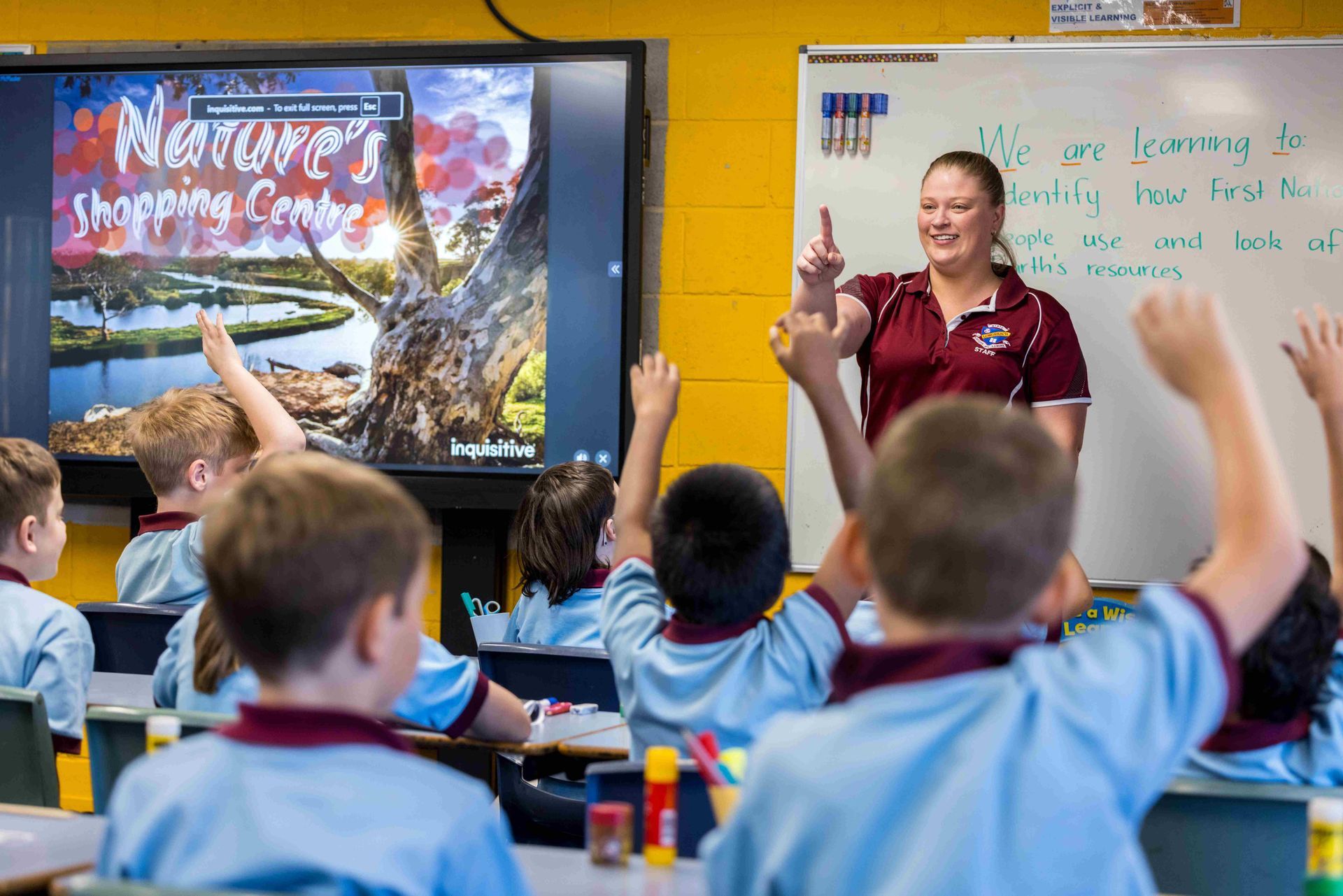 Teacher in classroom, pointing up at students with raised hands in front of interactive whiteboard.