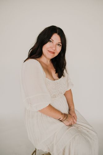A pregnant woman in a white dress is sitting on a stool.