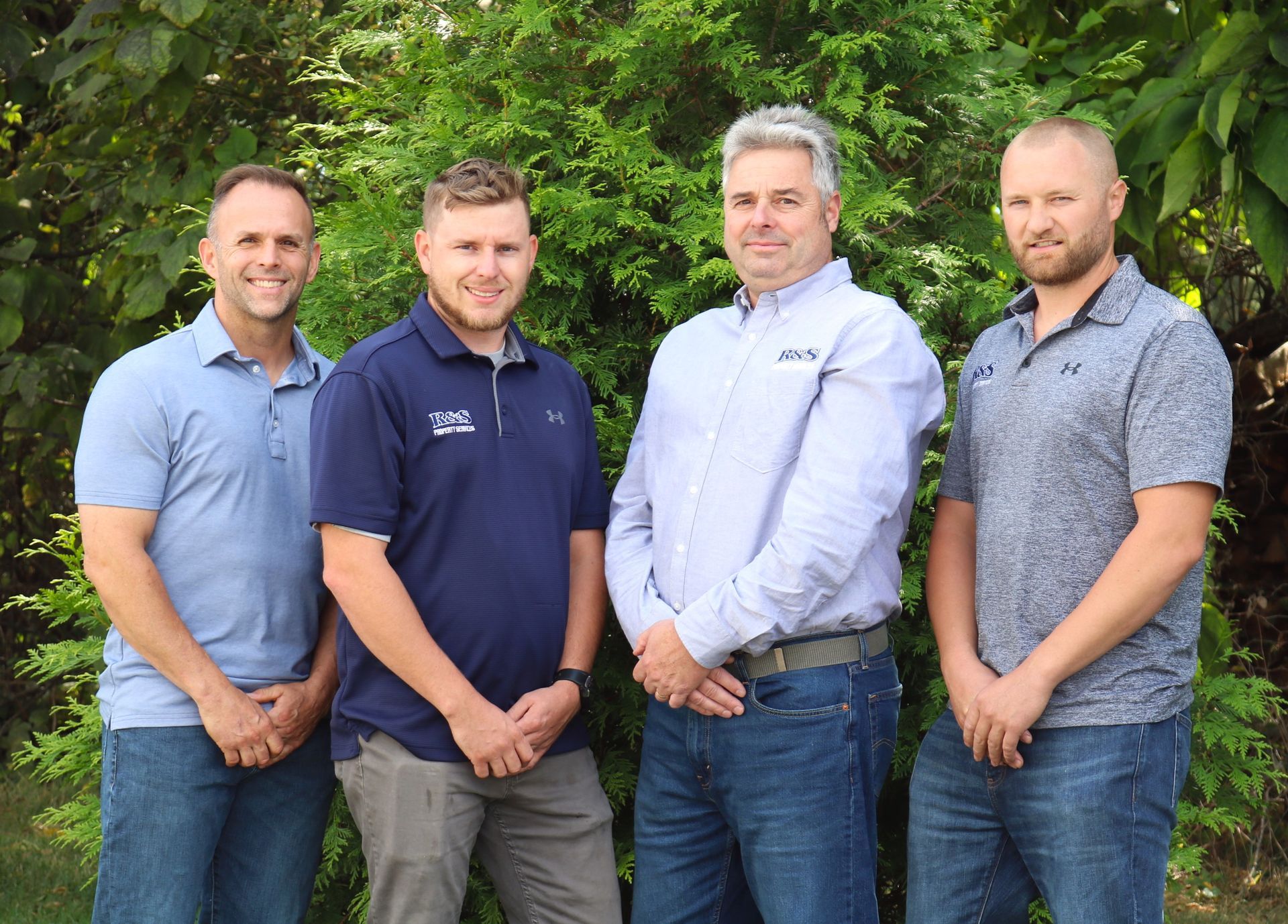 Four men standing in front of green bushes; they're wearing casual shirts and jeans.