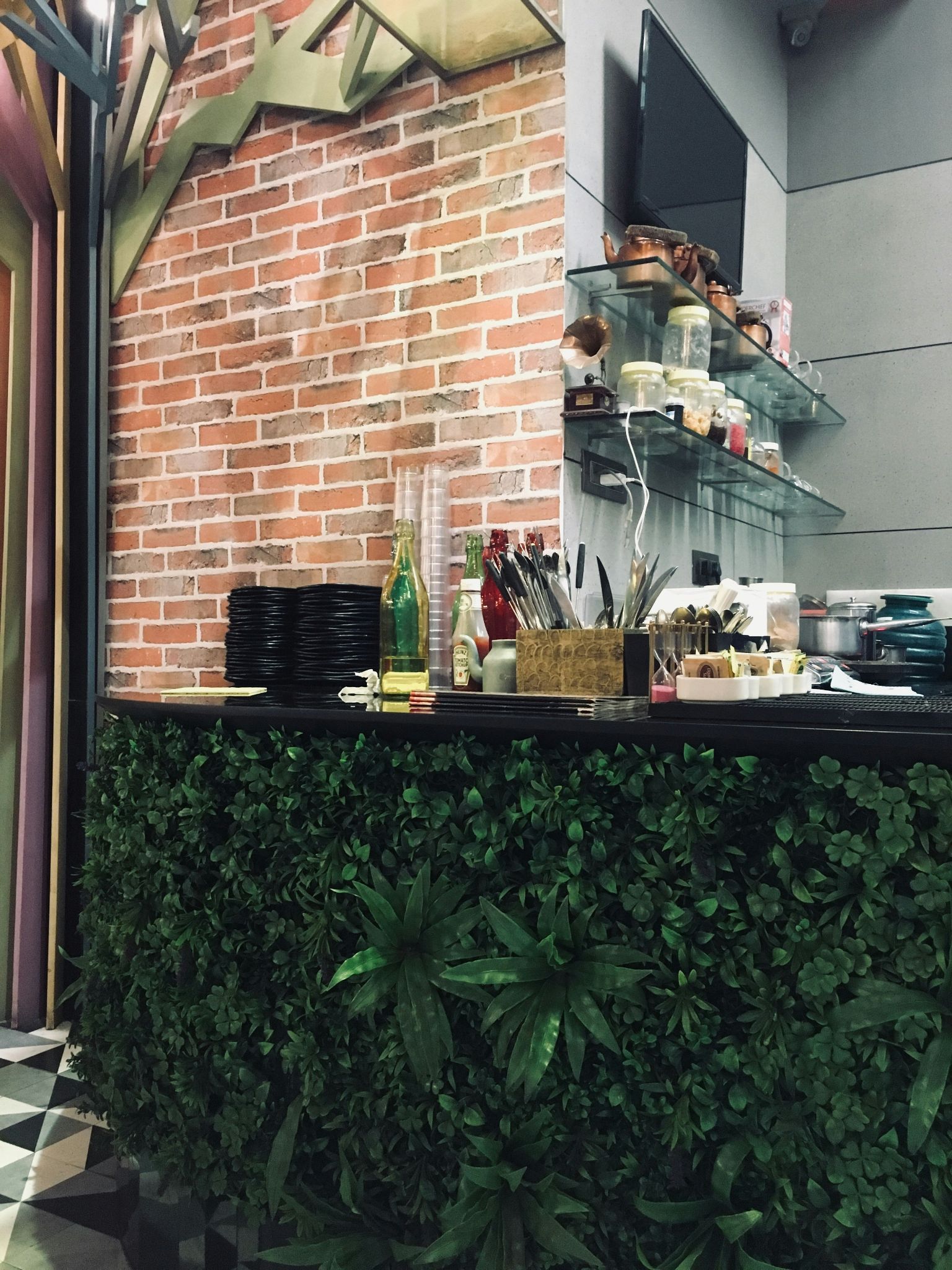 An outdoor kitchen counter full of greenery, utensils, plates and more. A TV is on the wall in the background above additional shelves.