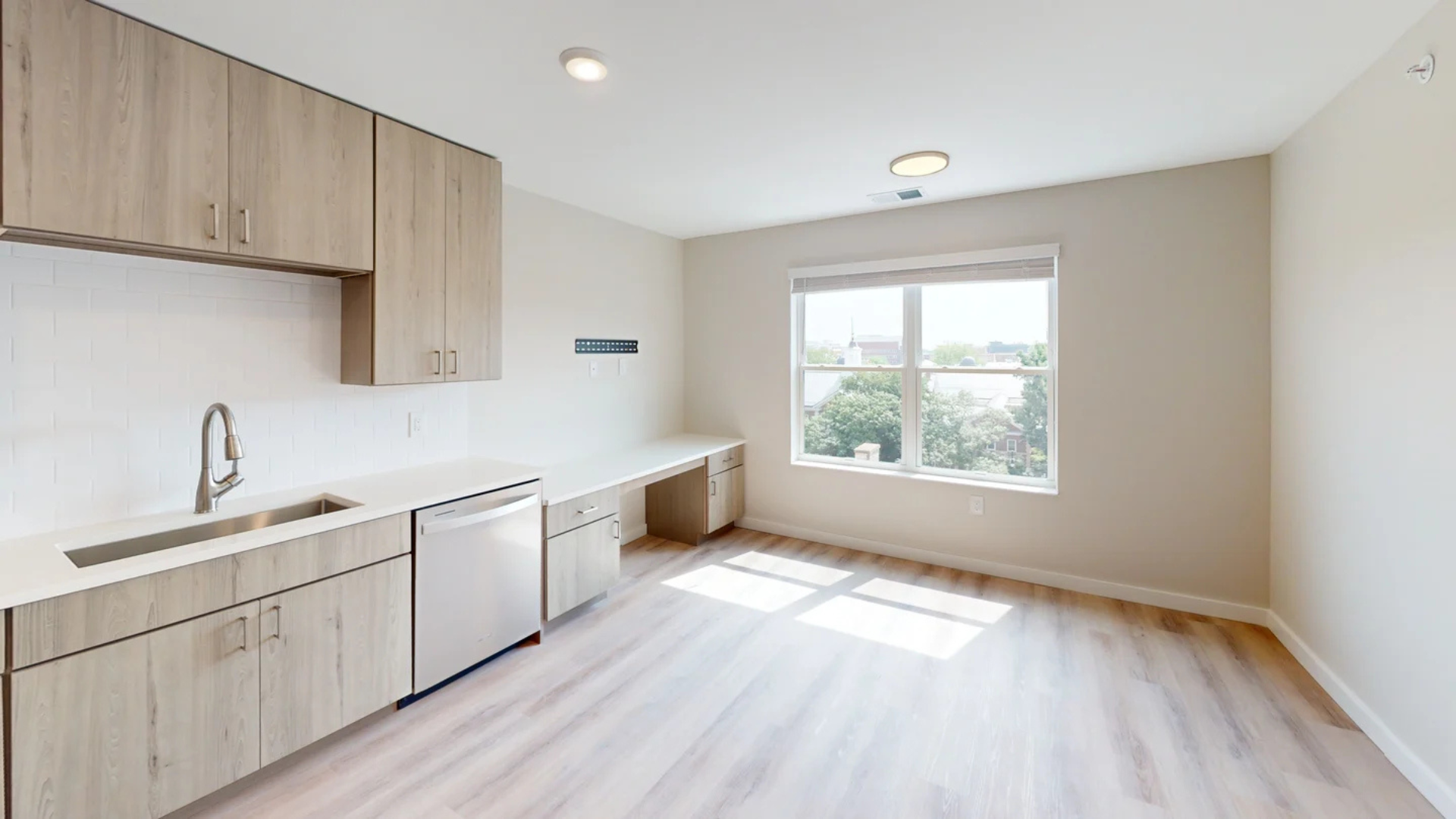 Bright, minimalist room with sink, cabinets, desk, and window. Light wood cabinets and floors.