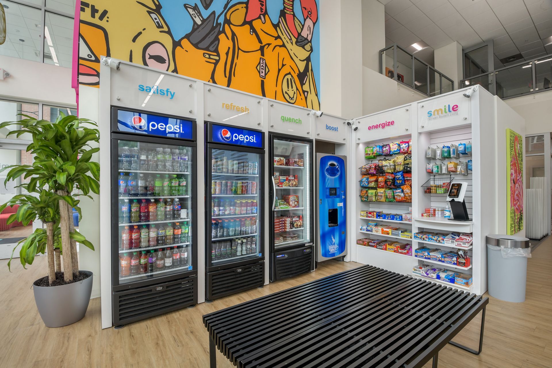 A well-lit room with vending machines. Coolers display Pepsi products and snacks, with a recycle machine in the middle. A black bench sits in front.