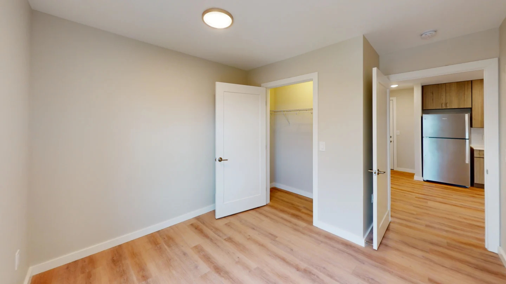 Empty bedroom with open closet and doorway to a kitchen area, with wood floors.