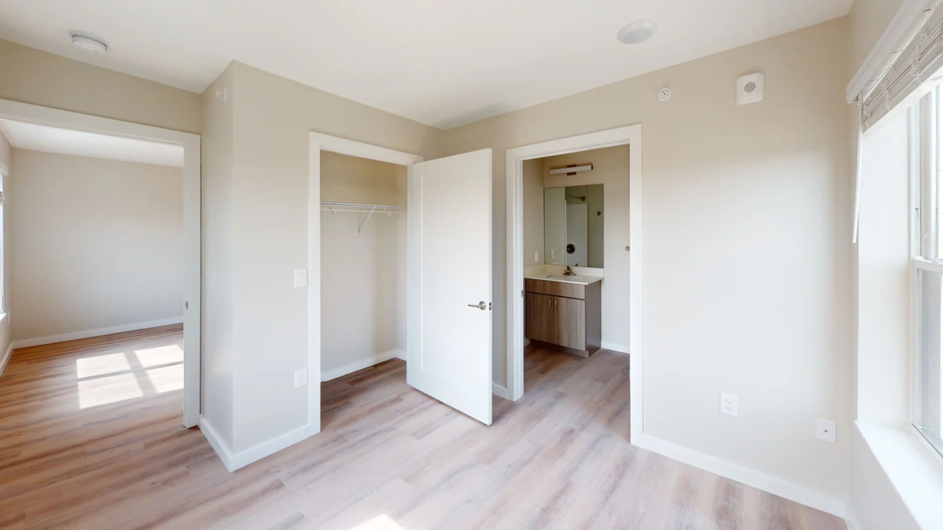 Empty bedroom with wood floors, closet, and adjoining bathroom. Natural light enters through the window.