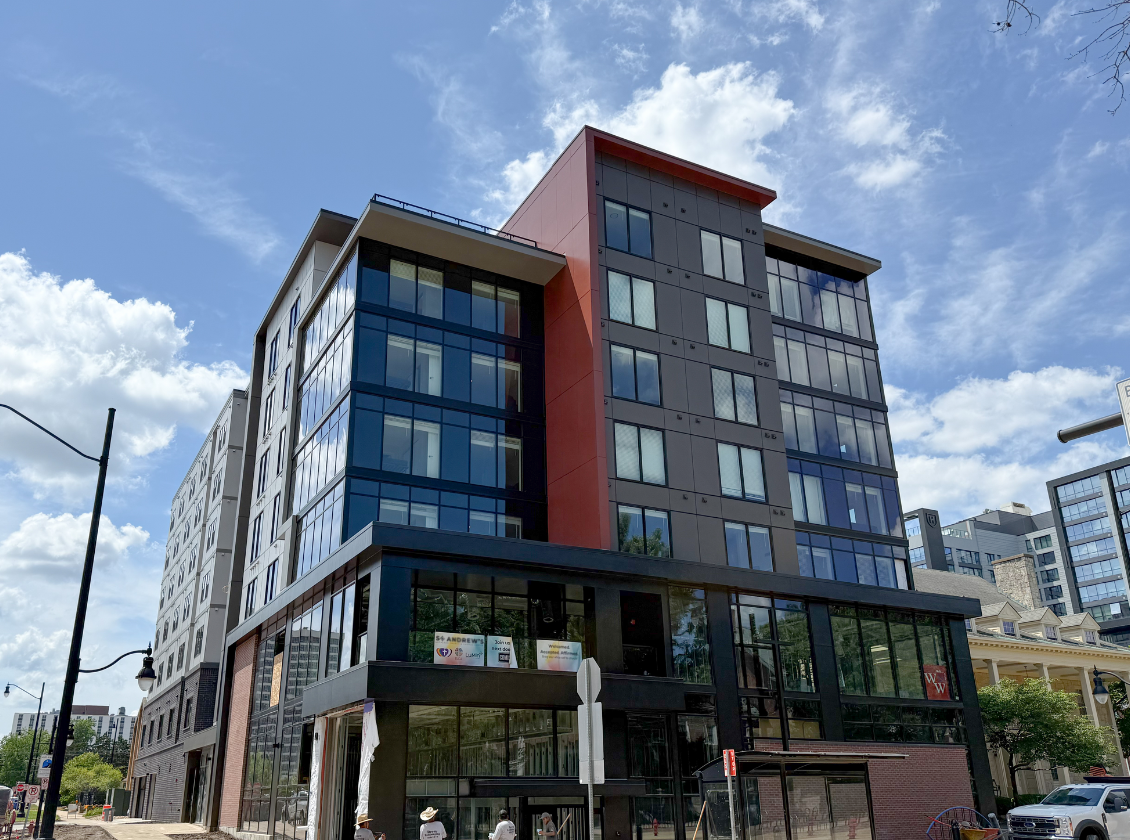 Modern multi-story building with dark gray and red accents; storefront on ground floor, sunny day.