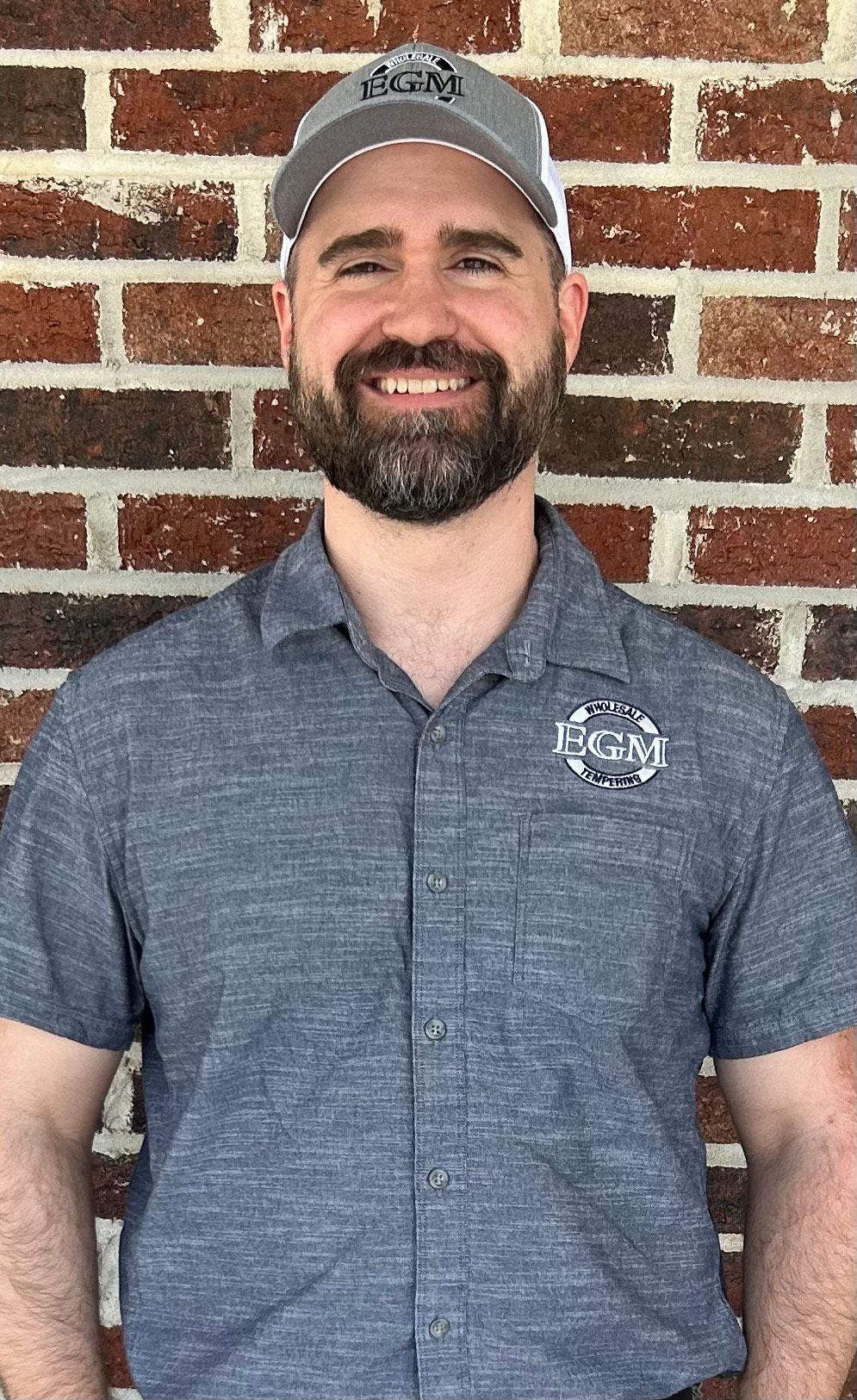 A man with a beard wearing a gray hat, short-sleeved shirt, and khaki pants stands in front of a brick wall.