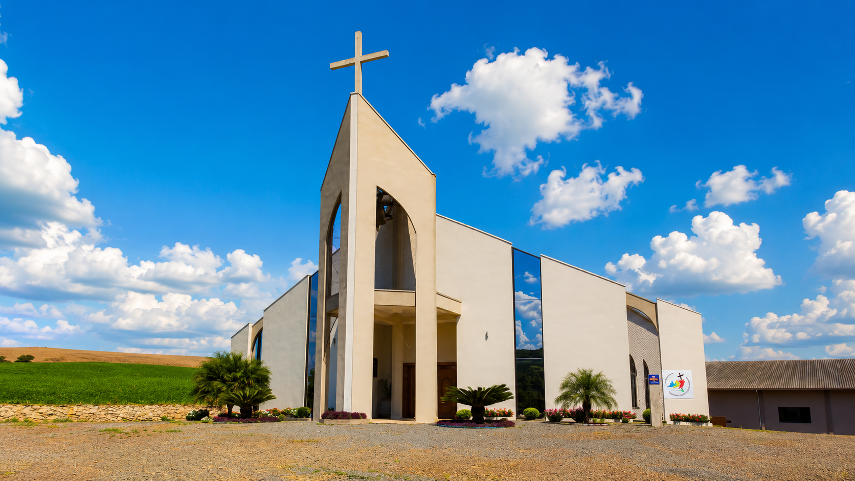 Igreja moderna branca com uma cruz e palmeiras sob um céu azul brilhante com nuvens.