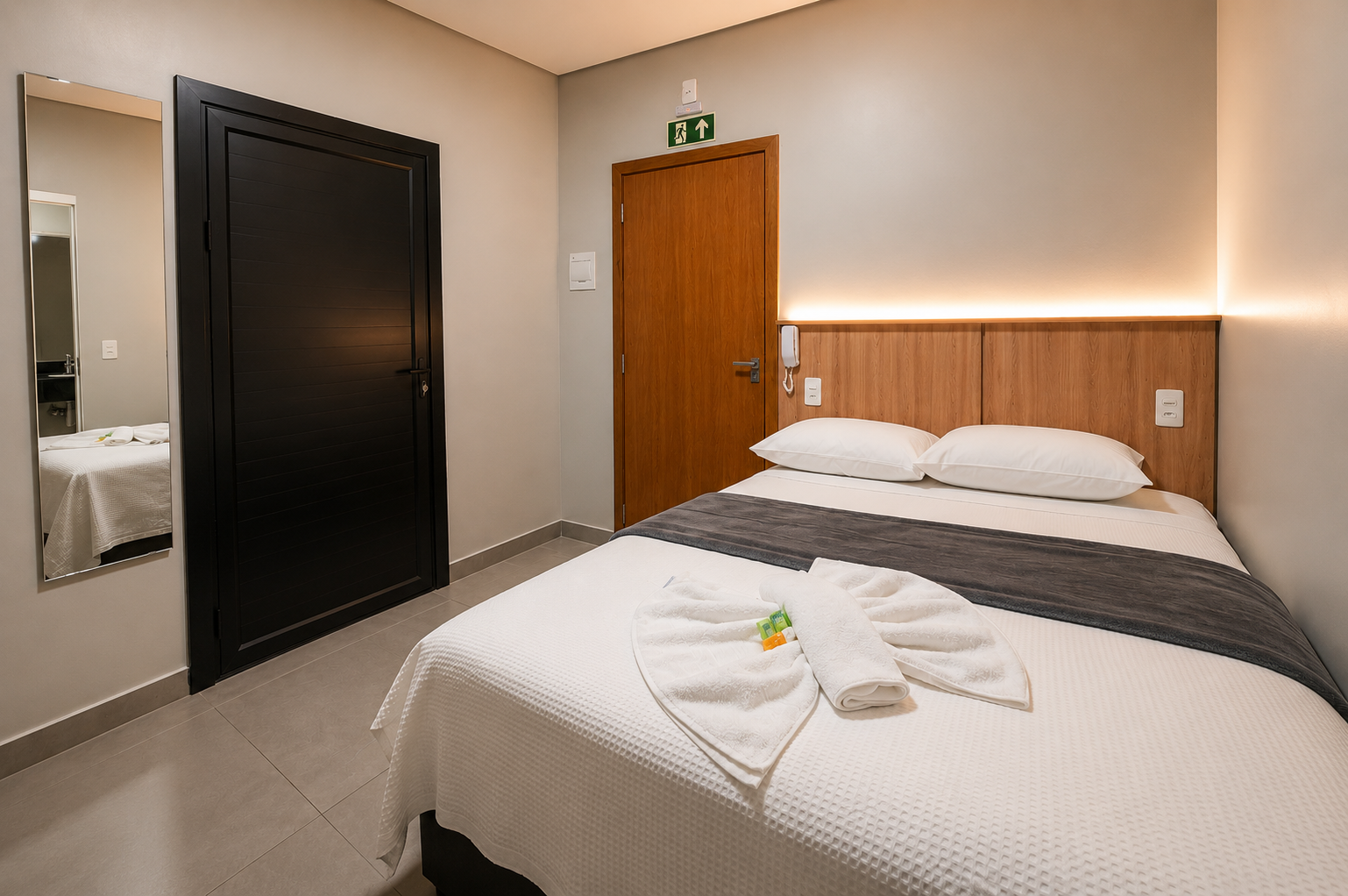Quarto de hotel compacto com duas camas, porta de madeira, guarda-roupa preto e pia com espelho na parede.