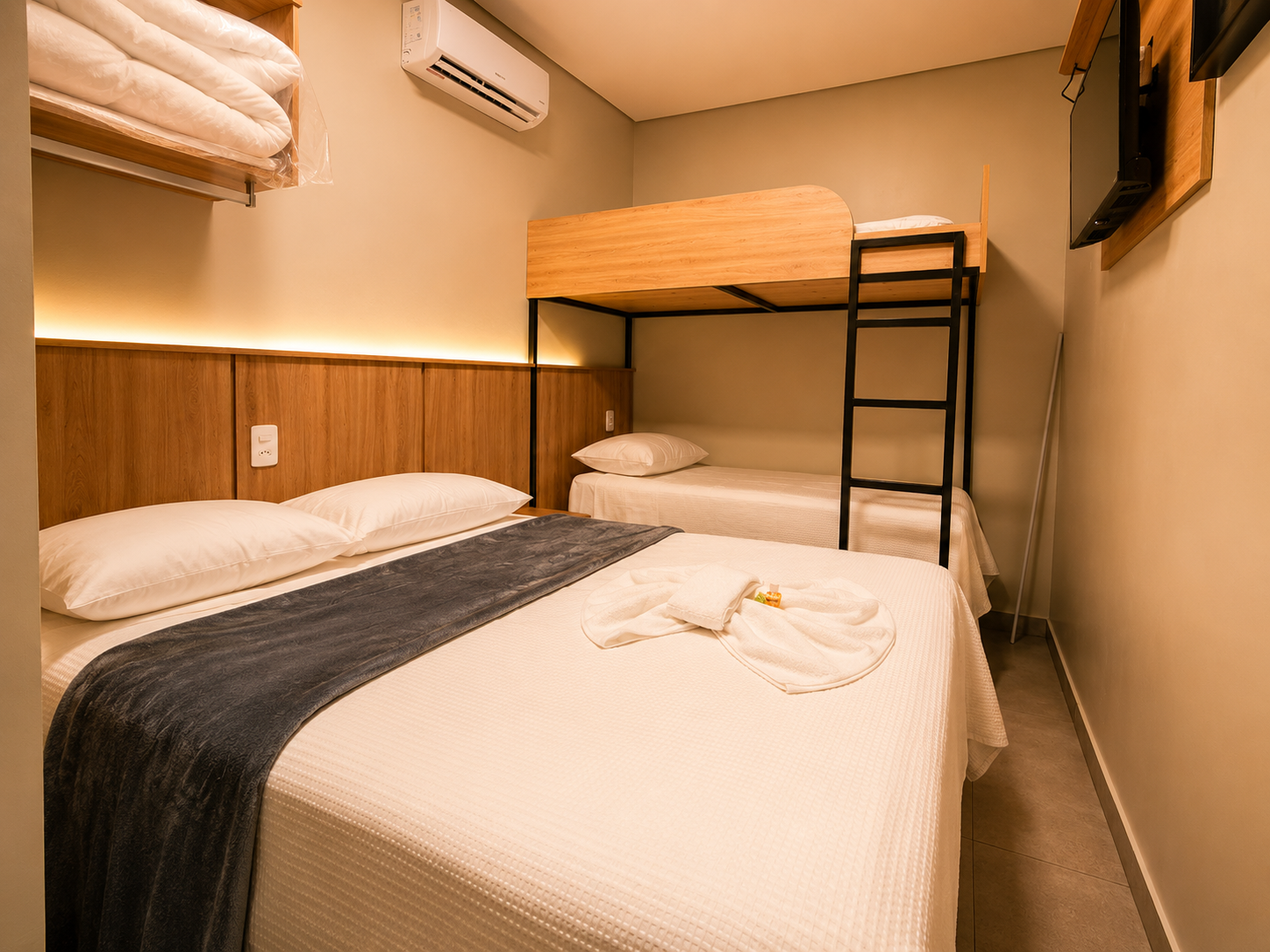 Quarto de hotel pequeno com duas camas, um beliche, lençóis brancos e painéis de madeira.