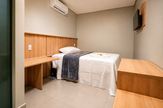 Quarto de hotel pequeno com duas camas, escrivaninhas de madeira, paredes bege e uma TV fixada na parede.