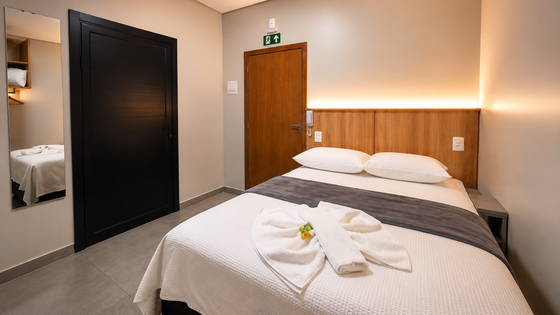 Quarto de hotel moderno com cama de casal, cabeceira de madeira, espelho e guarda-roupa preto.