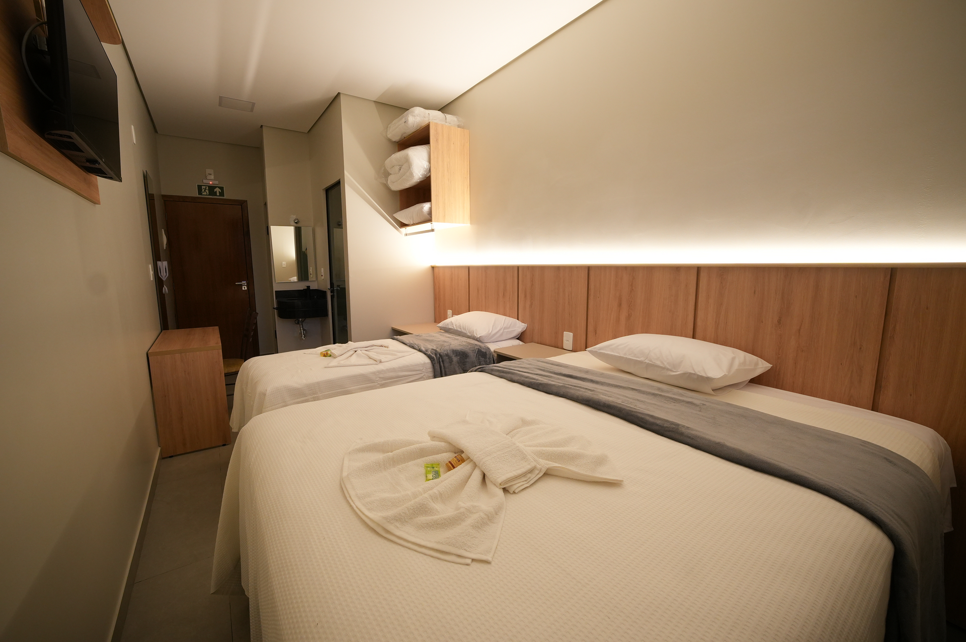 Quarto de hotel aconchegante com duas camas, painéis de madeira, iluminação suave e uma pequena prateleira junto à parede.