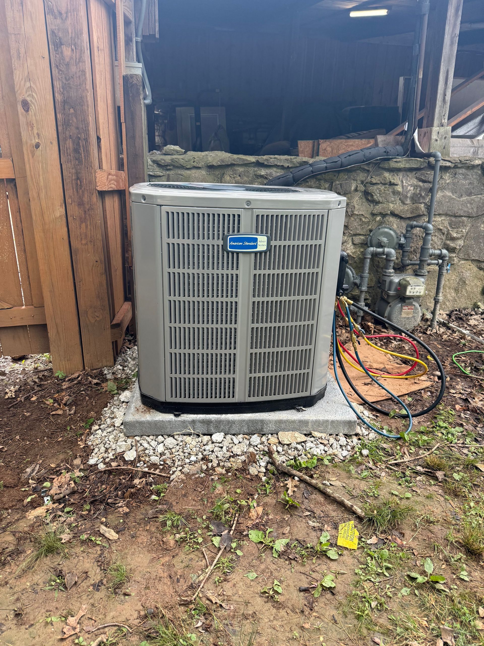 Outdoor HVAC condenser unit beside a stone wall, with hoses and debris around it