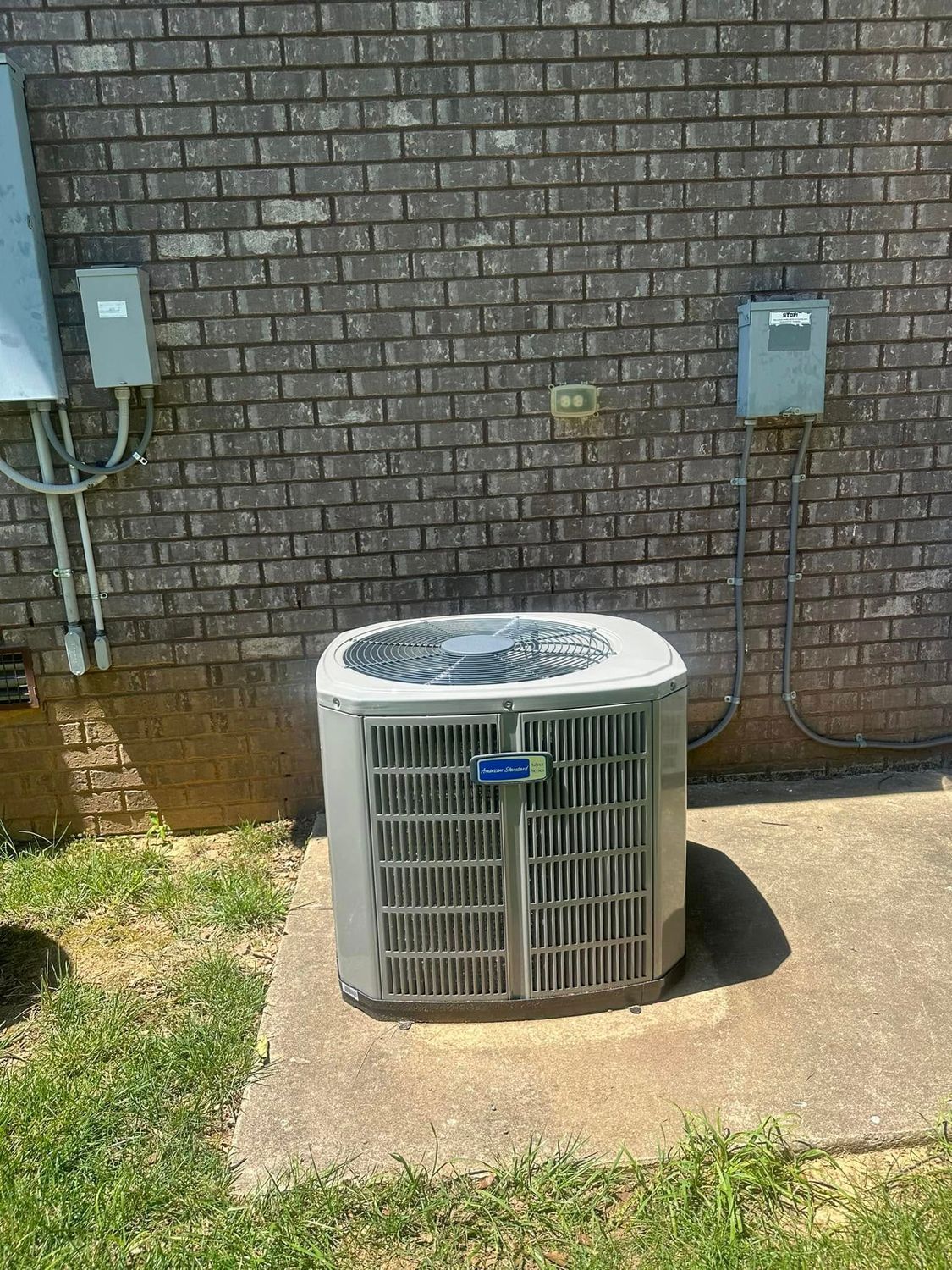 Outdoor air conditioner unit beside a brick wall with electrical boxes and a concrete pad.