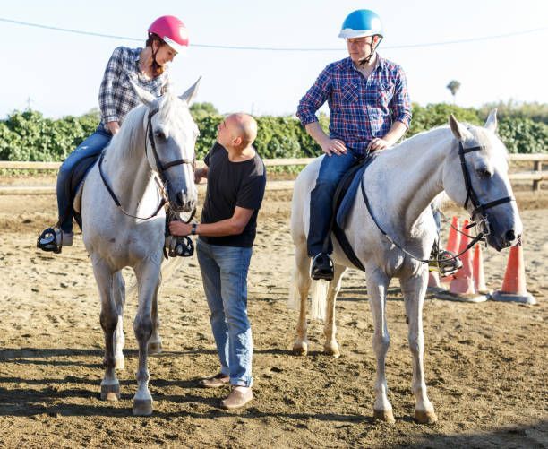 Two people on white horses with a bald man in jeans and t-shirt standing between them in an outdoor riding arena.