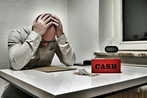 Una persona estresada sentada en un escritorio con la cabeza entre las manos, junto a una caja registradora roja y dinero esparcido.