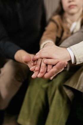 Manos apiladas en un gesto de apoyo grupal, con personas sentadas difuminadas al fondo.