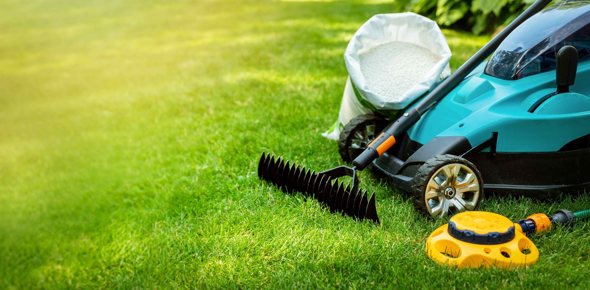 A teal lawnmower, a rake, and a yellow sprinkler sit on a bright green lawn beside a bag of fertilizer.