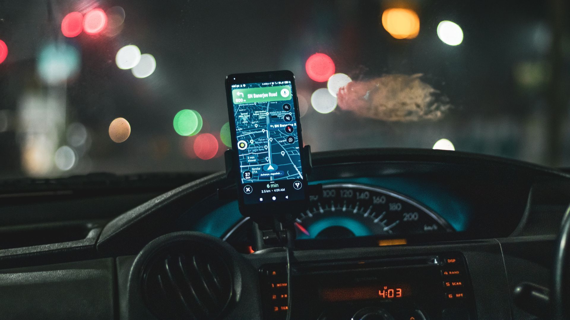 Phone mounted on car dashboard at night, displaying a map with blurred city lights ahead.