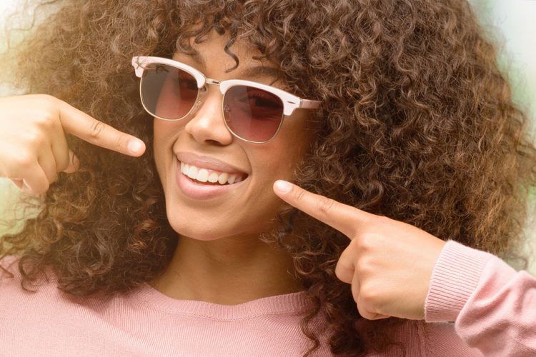 A woman with curly hair and sunglasses is smiling and pointing at her teeth.