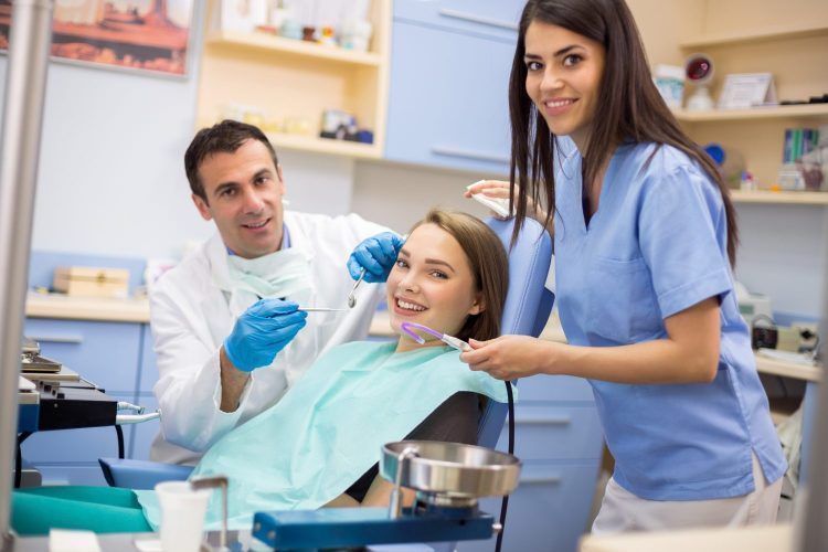 A woman is sitting in a dental chair while two dentists are working on her teeth.