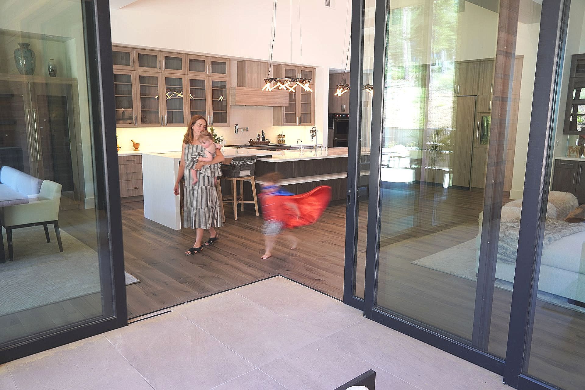 Woman holding baby and watching child run through sliding glass door.