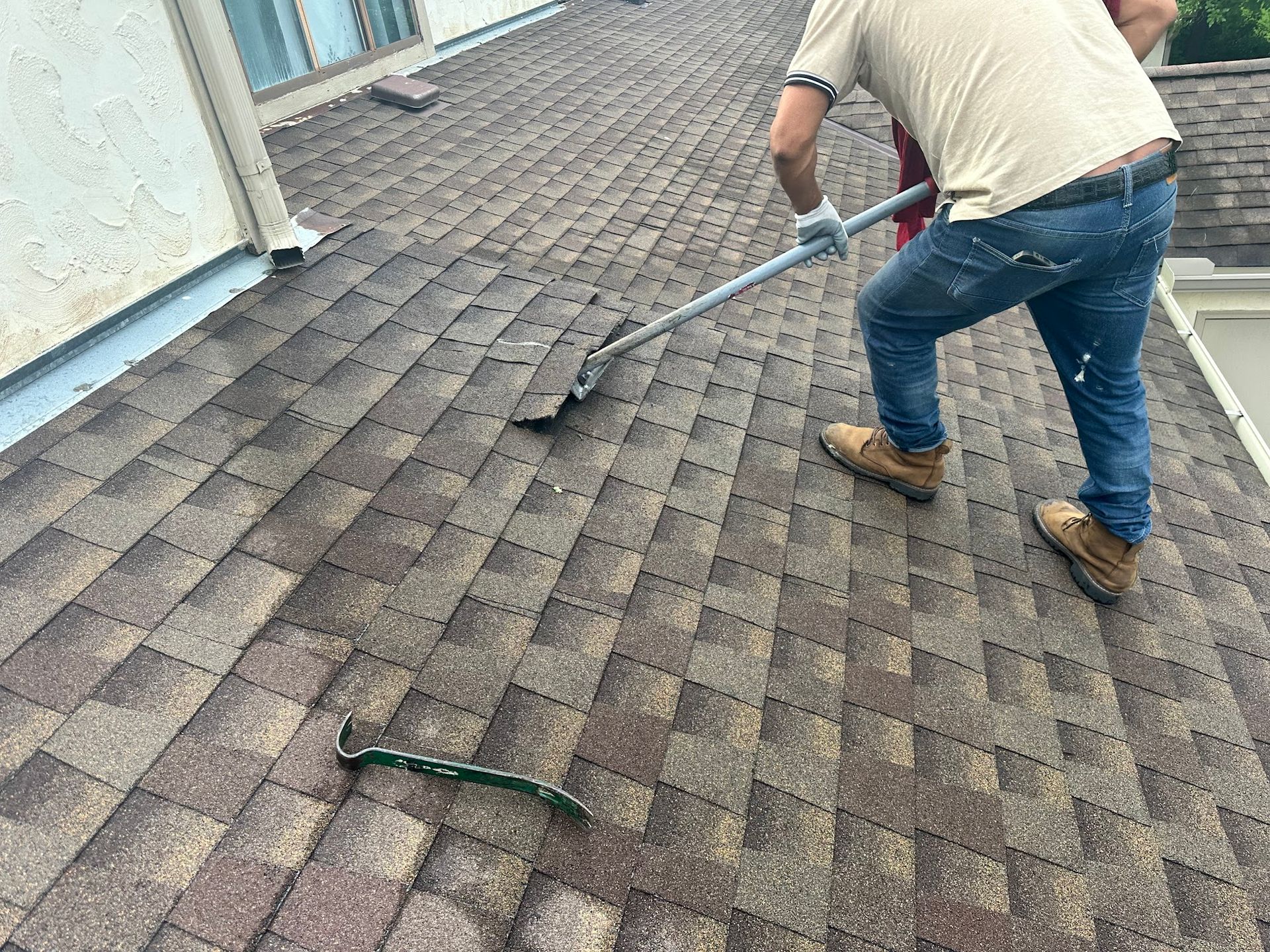 Man on a roof using a long-handled tool to sweep debris. Brown shingles, blue jeans, tan shirt.