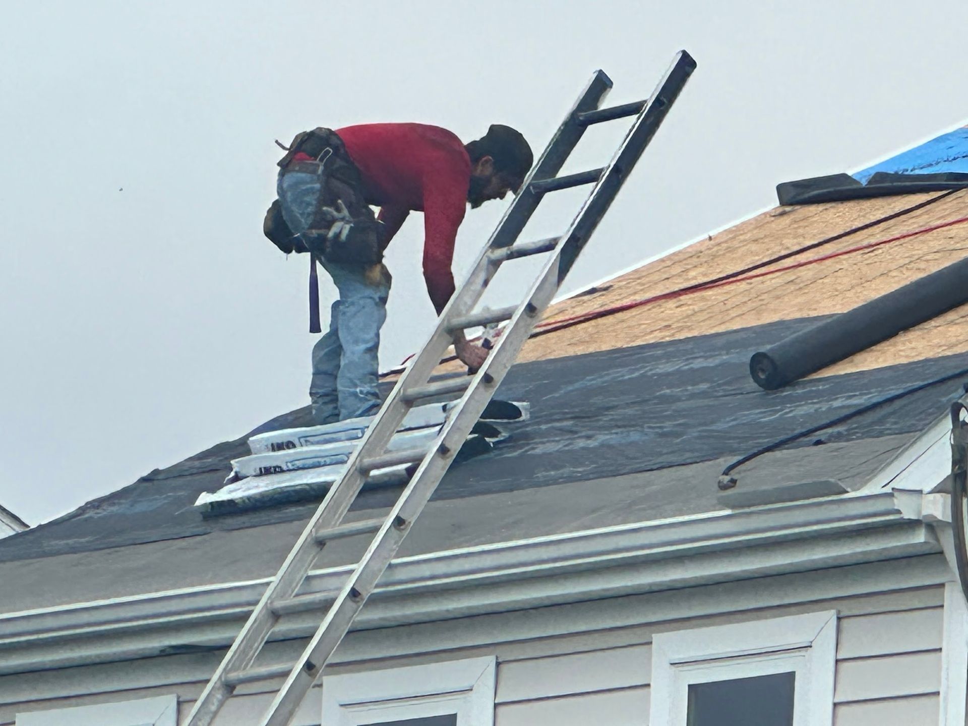 Roofer in a red shirt on a ladder, working on a roof with shingles.