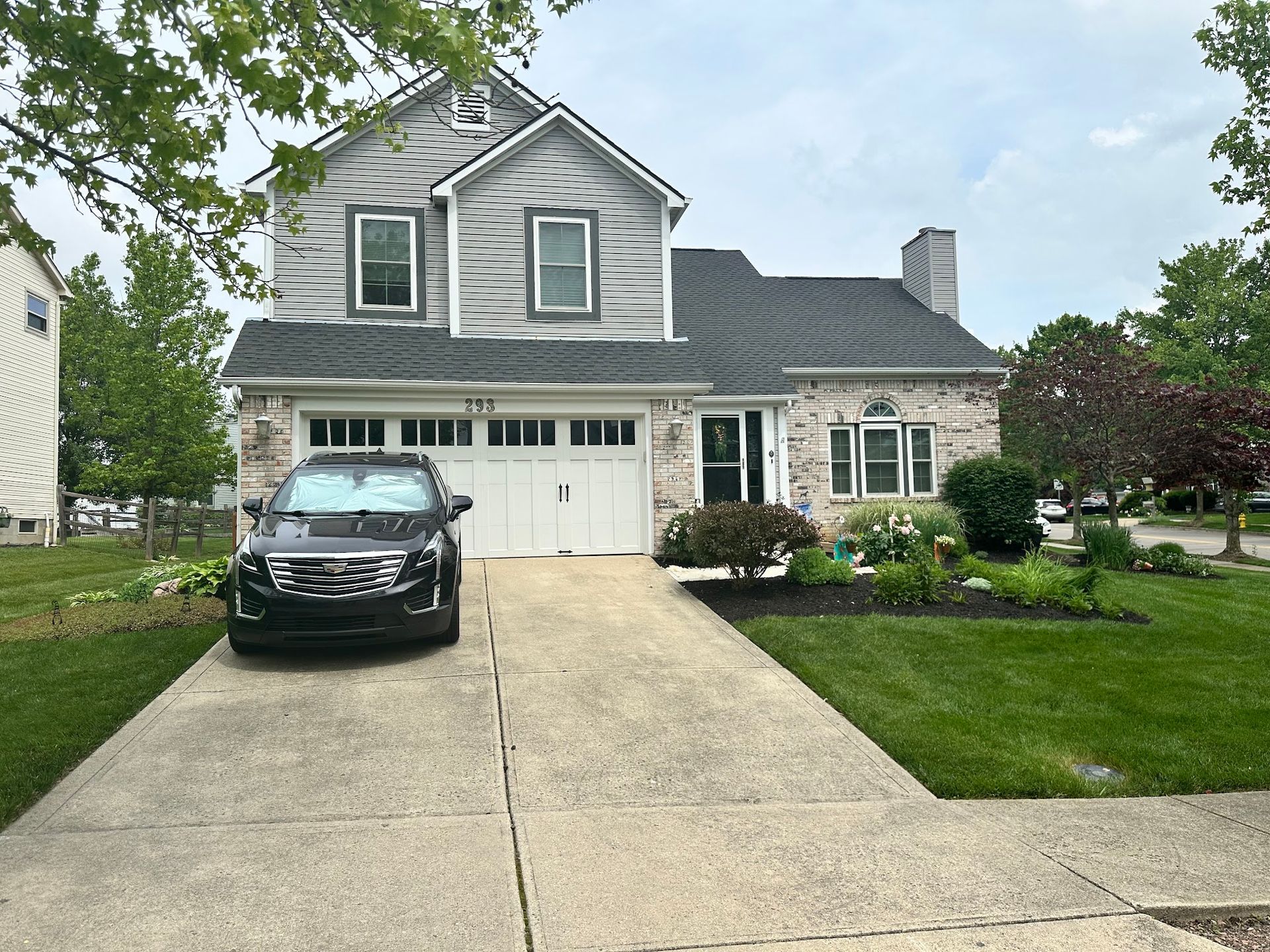 Two-story house with gray siding and brick facade, white garage door, and a black car parked in the driveway.
