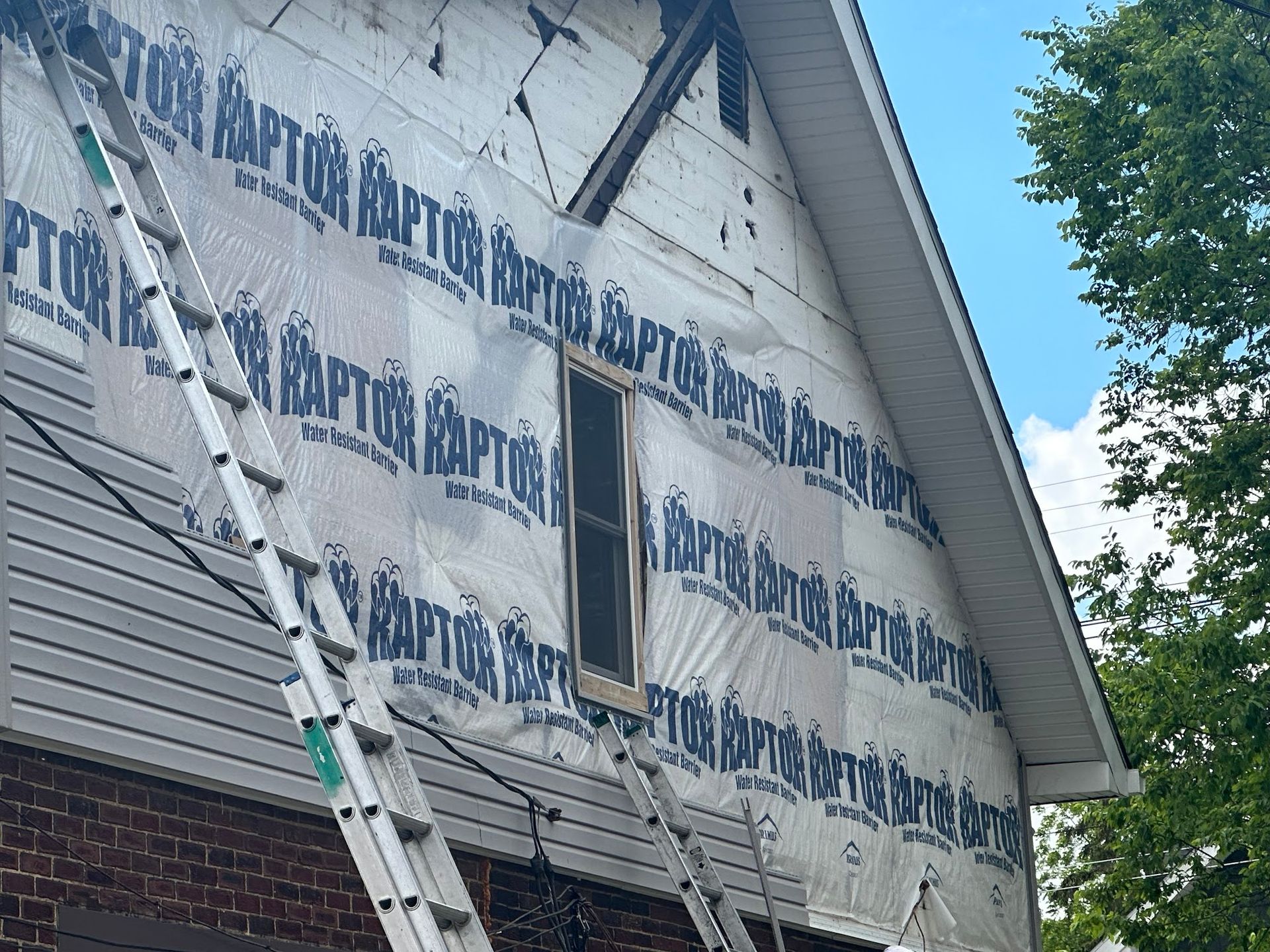House with blue wrap, siding, and ladders. Construction in progress against a blue sky.