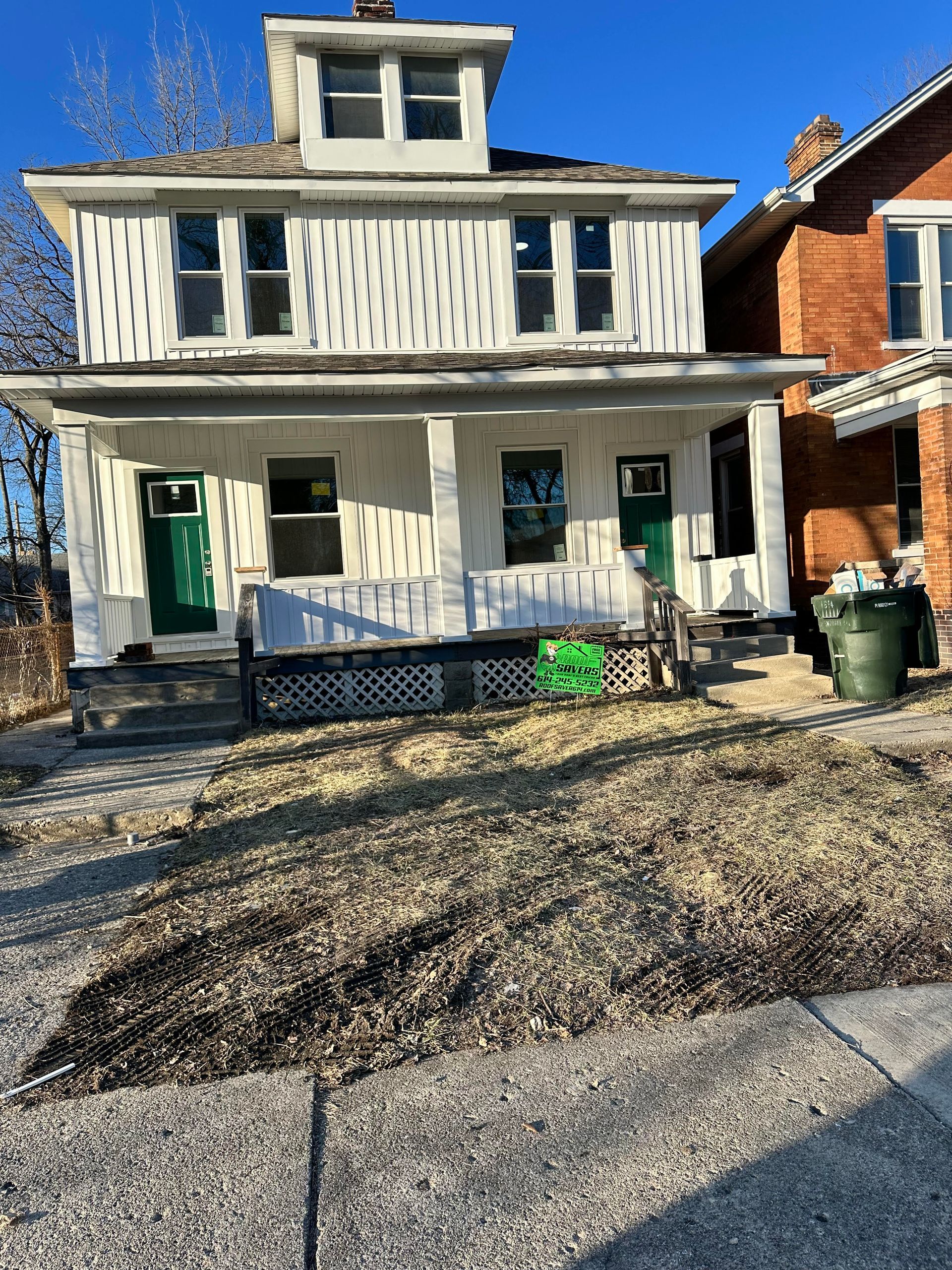 Two-story house with white siding, green doors, and a porch. Brown grass in front, and a brick house to the right.