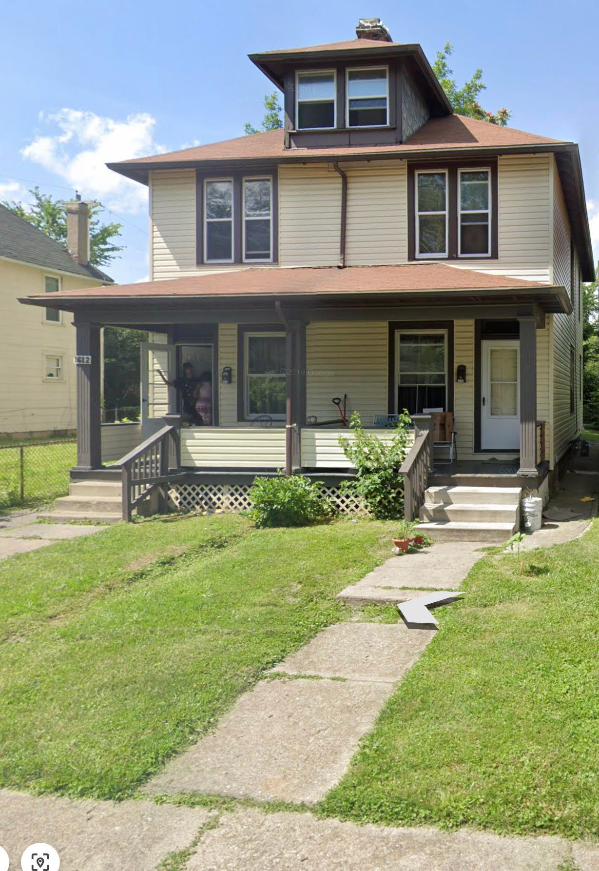 Two-story tan house with porch and dormer. Green lawn and sidewalk in front.
