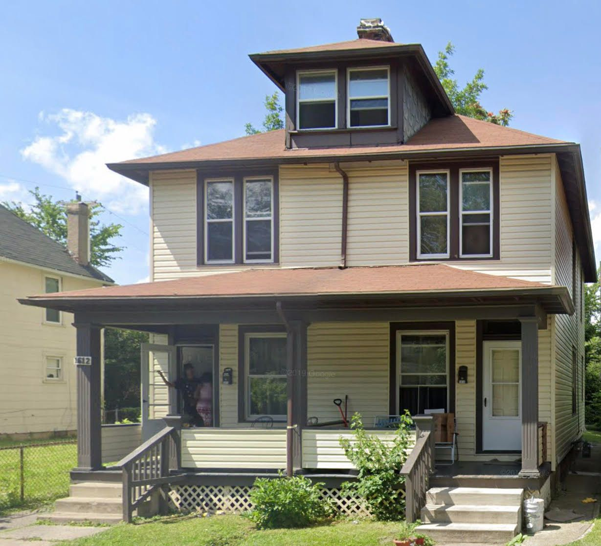 Two-story house with porch, tan siding, brown trim, and a dormer. Blue sky.