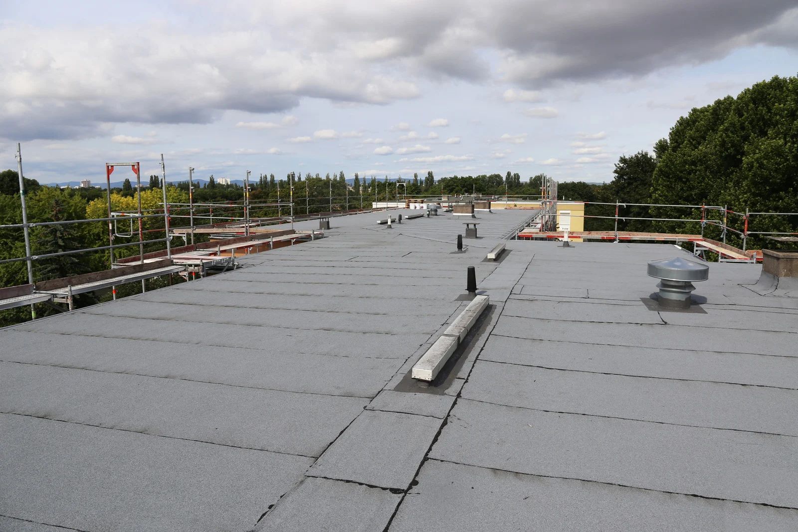 Flat, gray roof with ventilation, surrounded by scaffolding and trees under a cloudy sky.