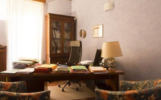 Scrivania da ufficio piena di documenti e fascicoli, con sedia e lampada. Mobiletto in legno con libri.