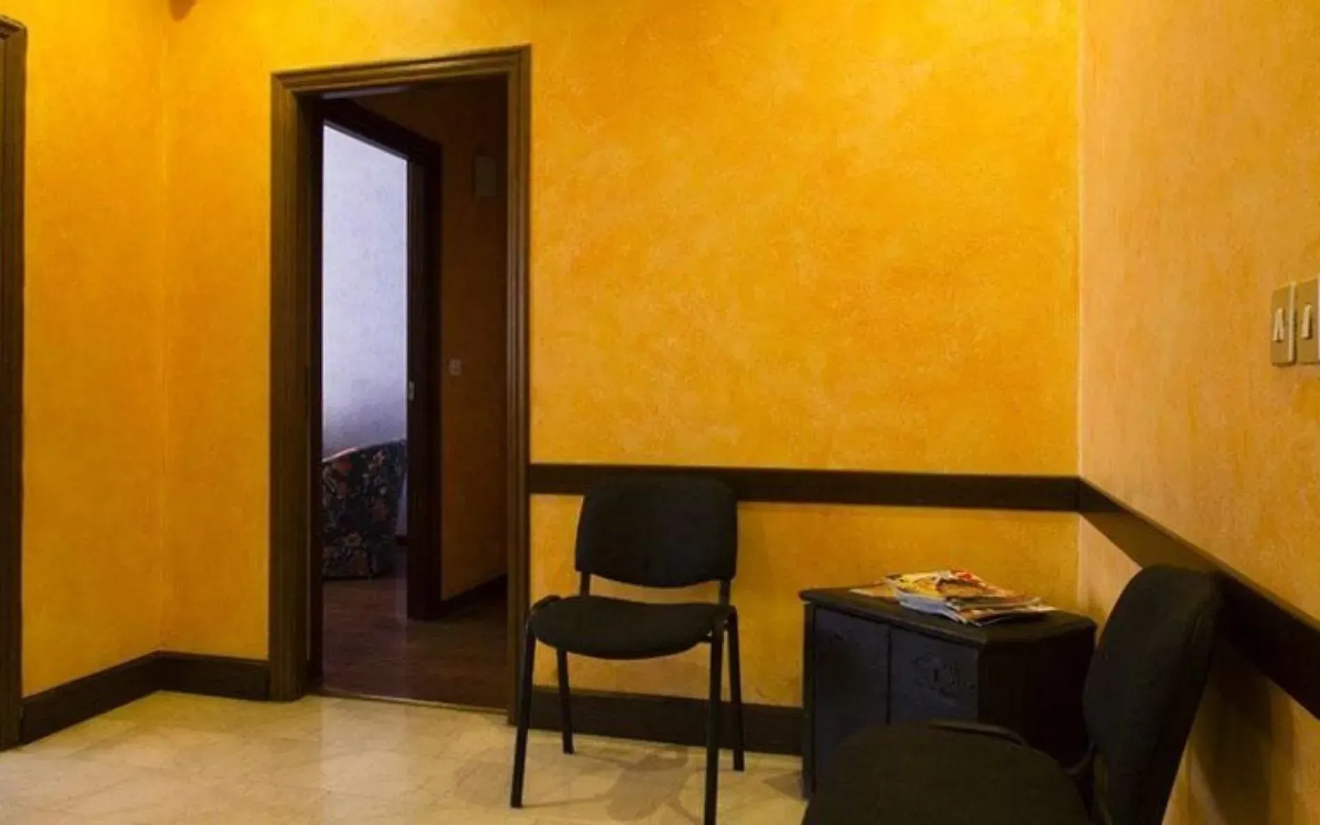 Sala d'attesa con pareti gialle, finiture scure, sedie nere e una porta.