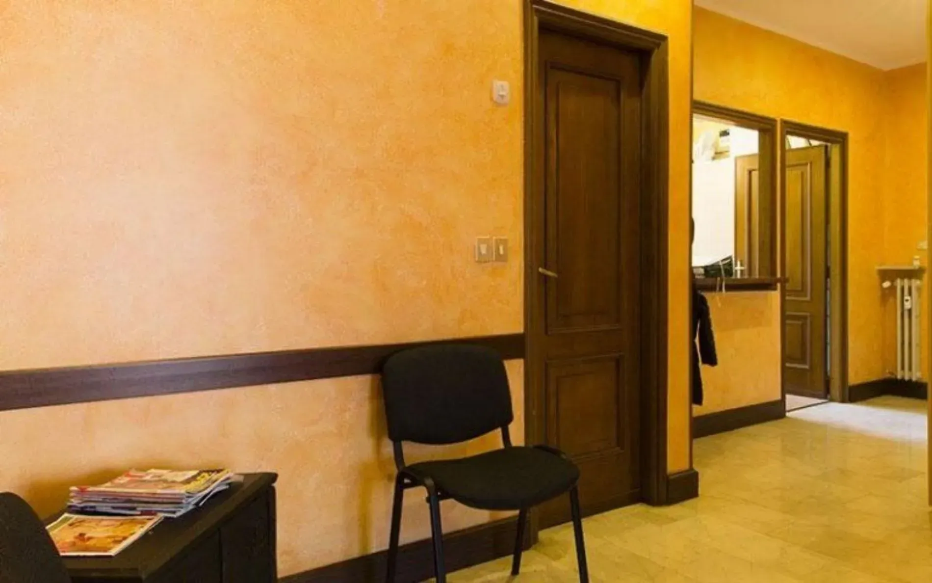 Sala d'attesa con pareti arancioni, porte marroni, una sedia nera e riviste su un tavolo.
