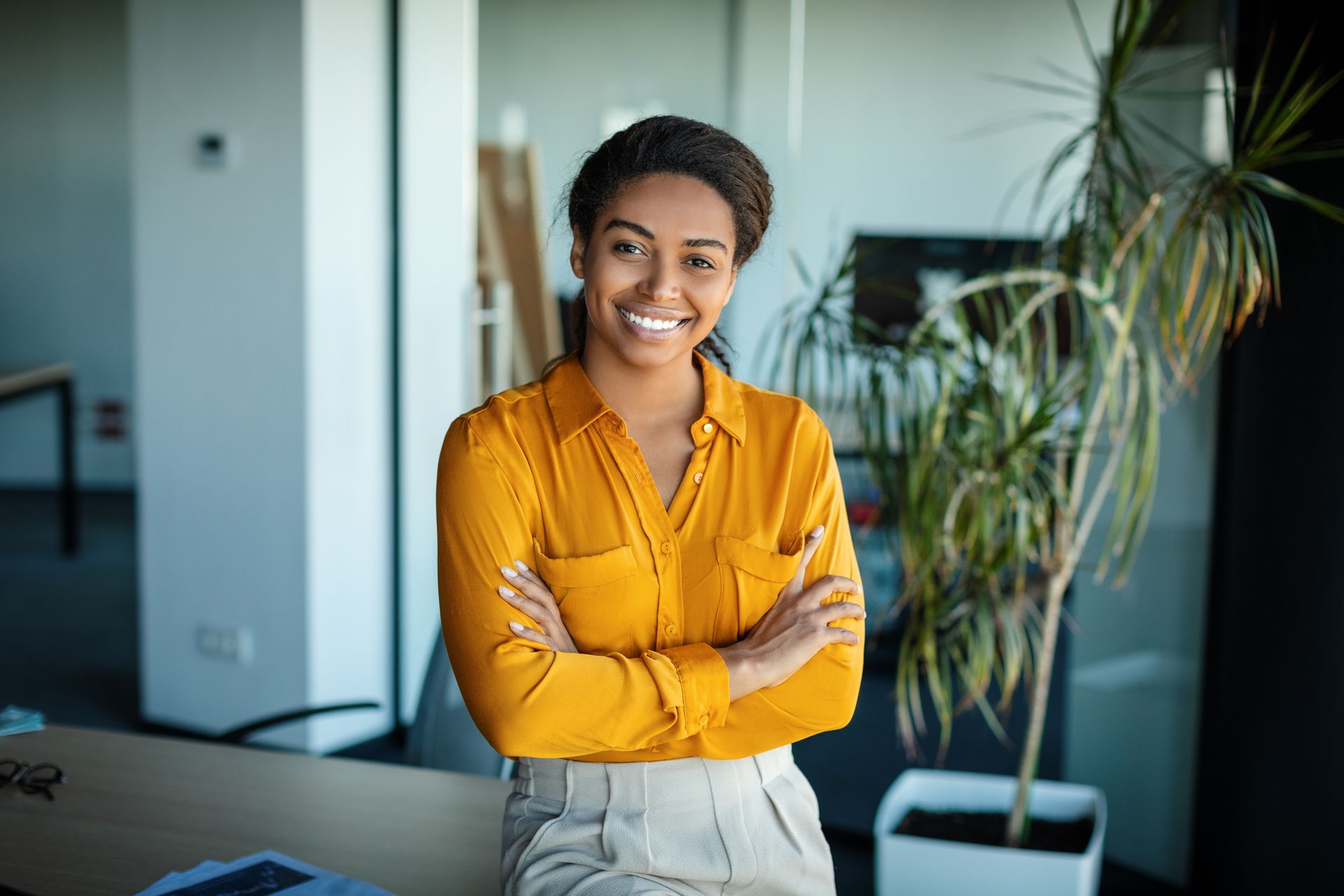 Smiling person in a mustard blouse stands with arms crossed in a bright office.