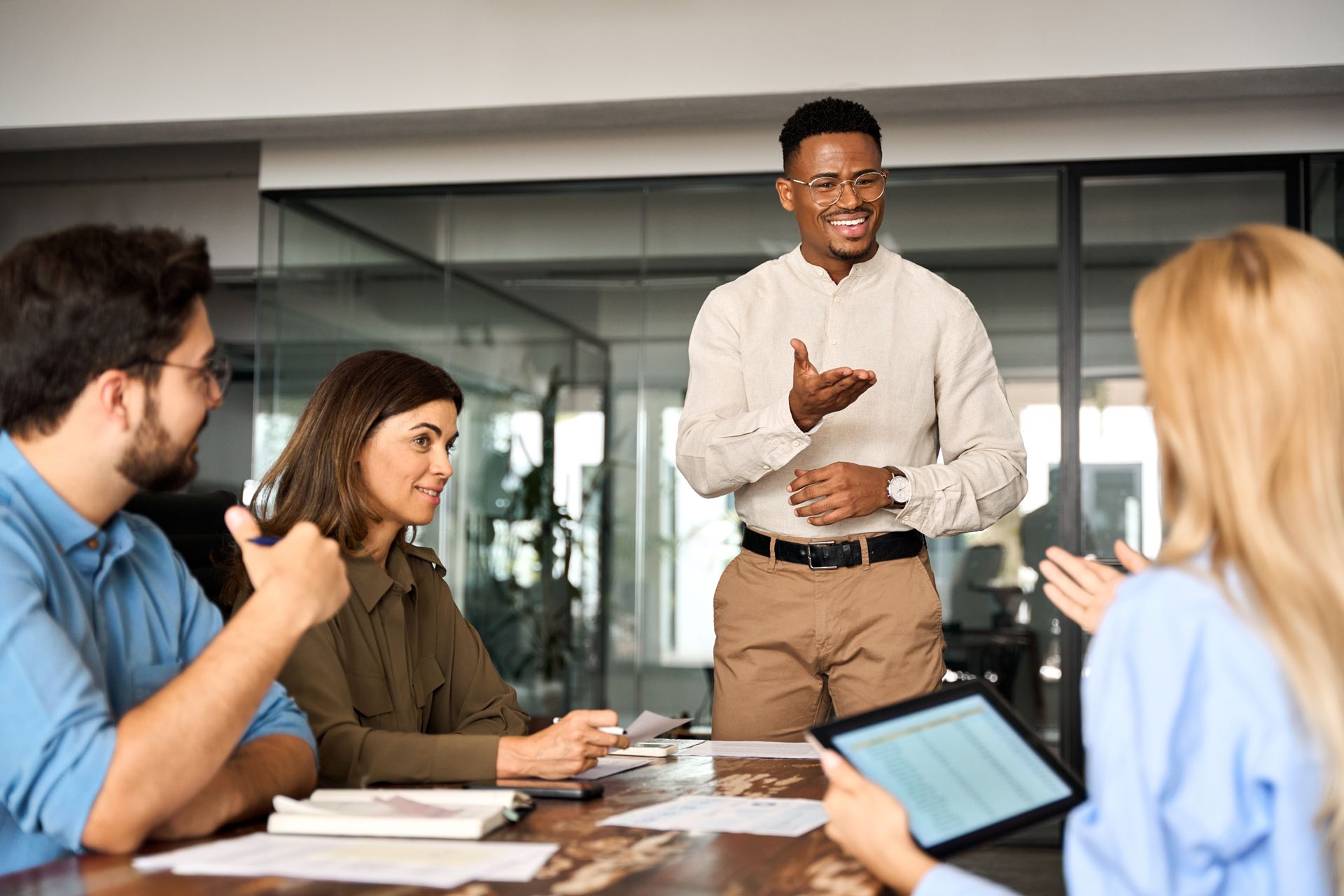 Team meeting in a glass-walled conference room, with one person presenting while others listen and take notes.