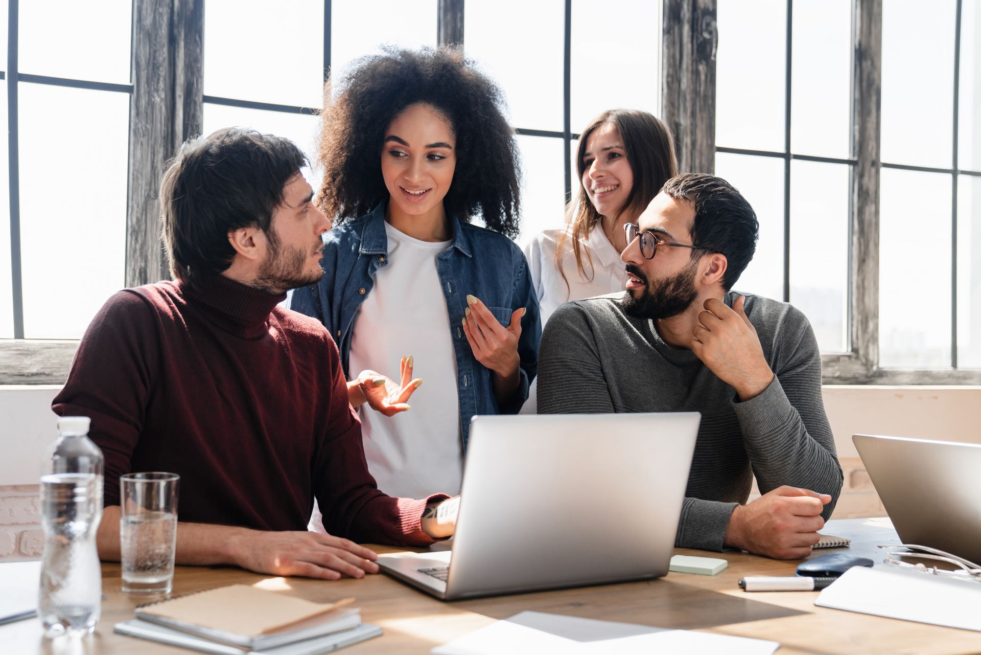 Four coworkers discussing around a laptop at a bright office table, one gesturing while others listen