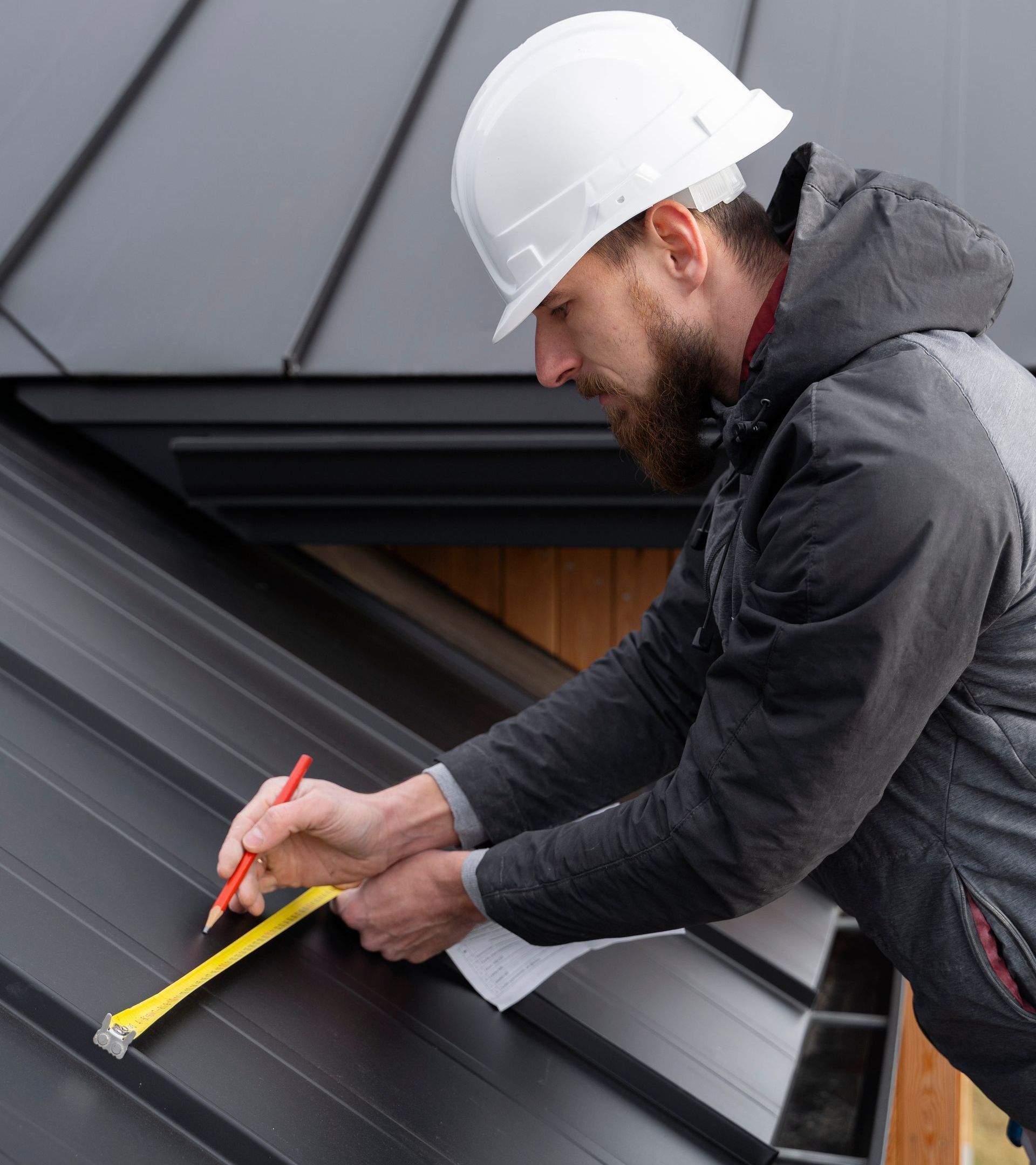 A man wearing a hard hat is measuring a roof with a tape measure