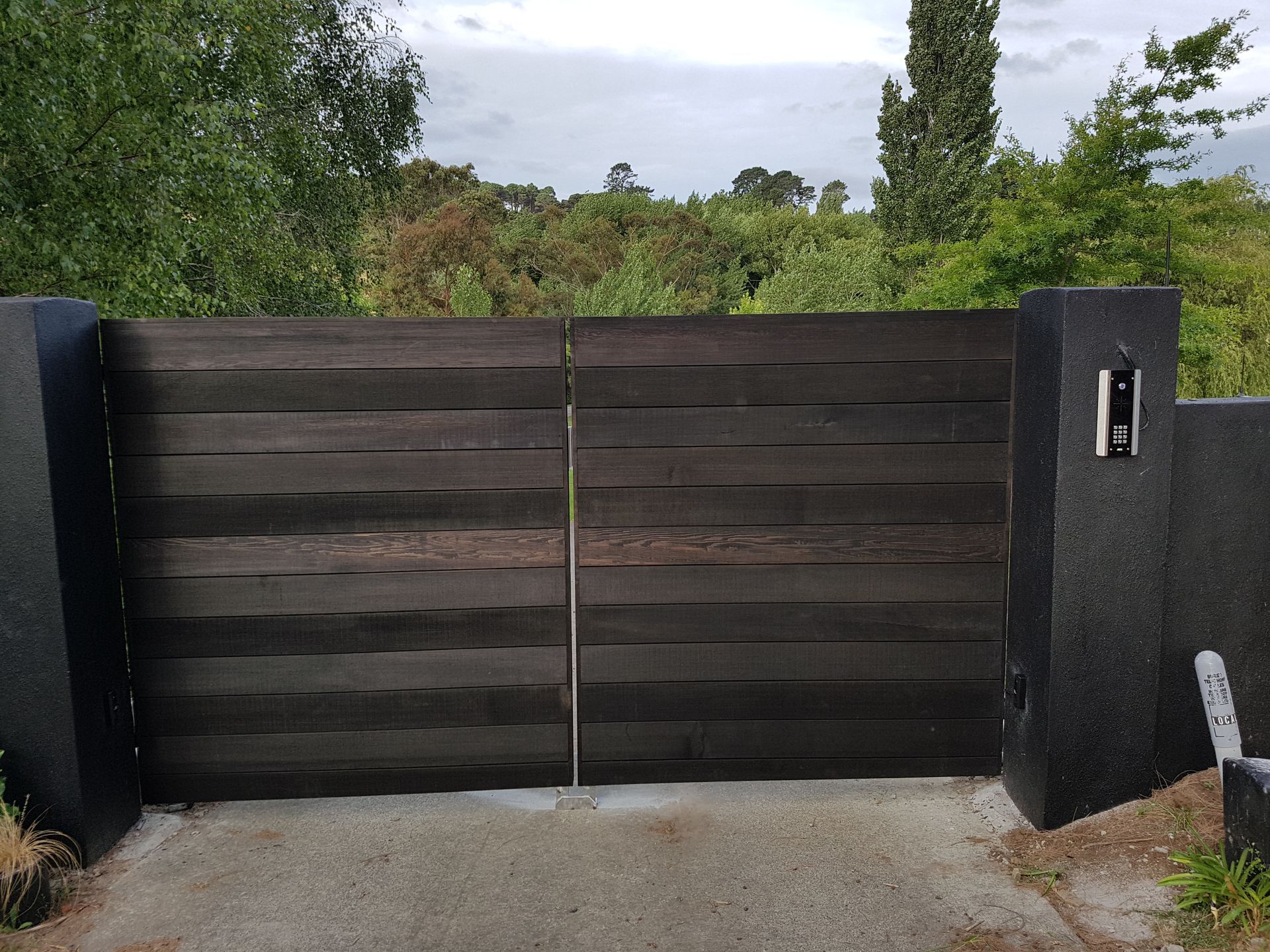 A wooden gate is open to a driveway with trees in the background